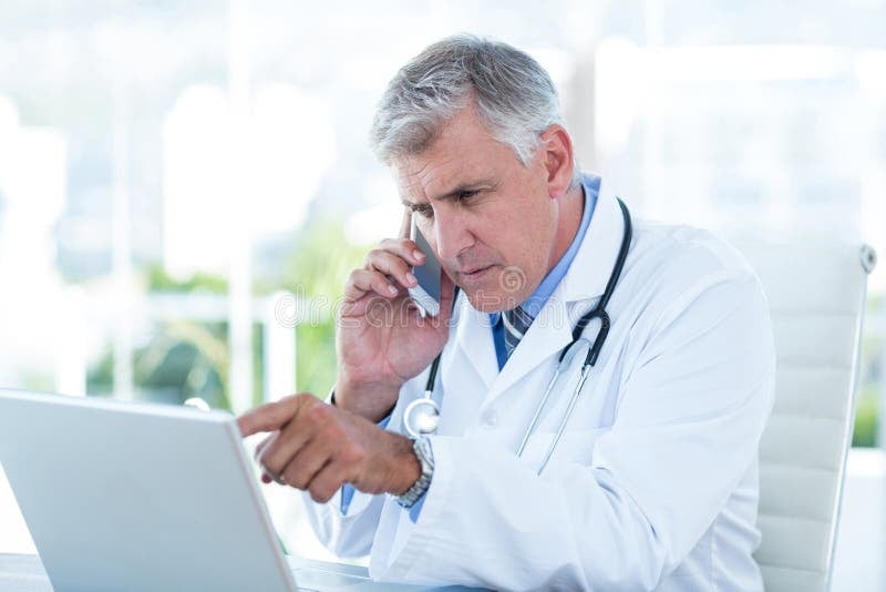 serious-doctor-working-laptop-having-phone-call-medical-office-54763710